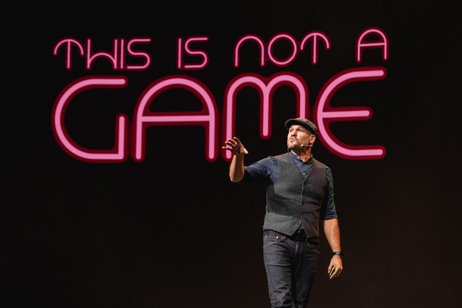 Alex on stage in front of a neon 'This is not a game' backdrop